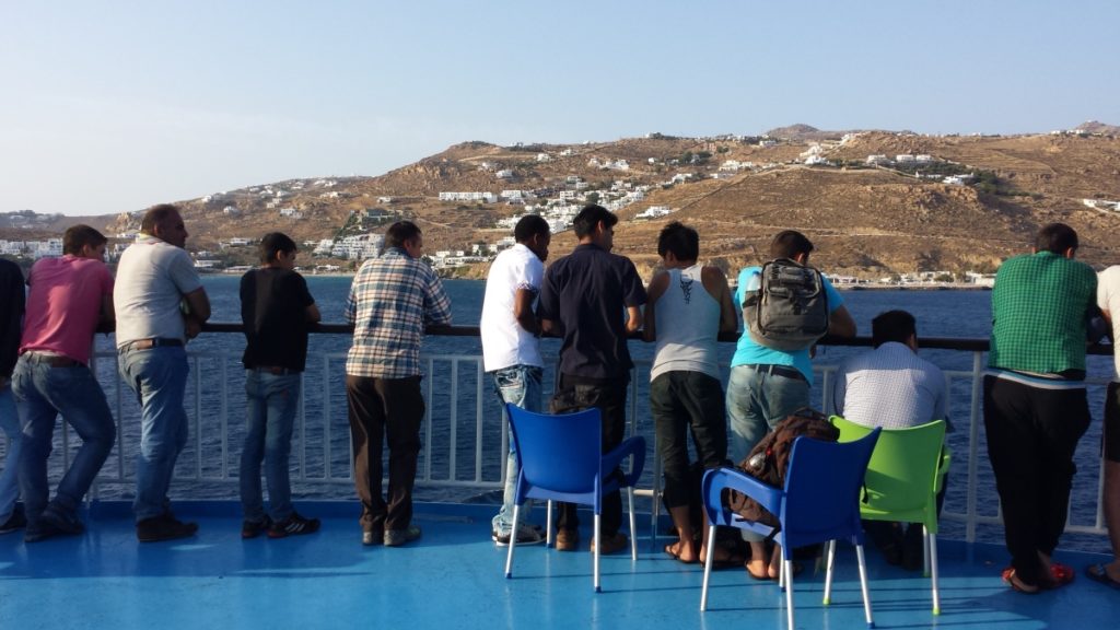 Männer stehen an der Reeling. Dahinter ist die Insel Mykonos zu sehen.
