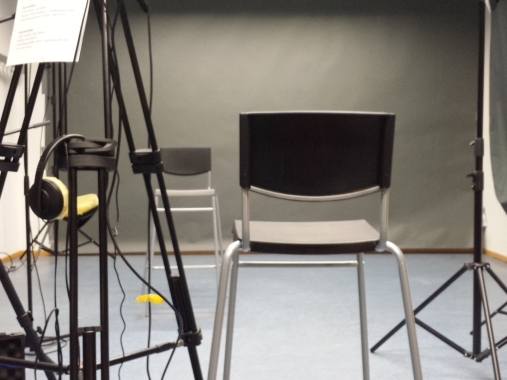 Studiosituation: Zwei Stühle umgeben von Licht- und Kamerastativen vor grauer Wand