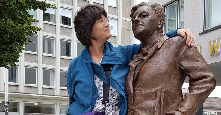 Frau mit blau-türkiser Jacke hat ihren Arm bewundernd um eine Statue gelegt